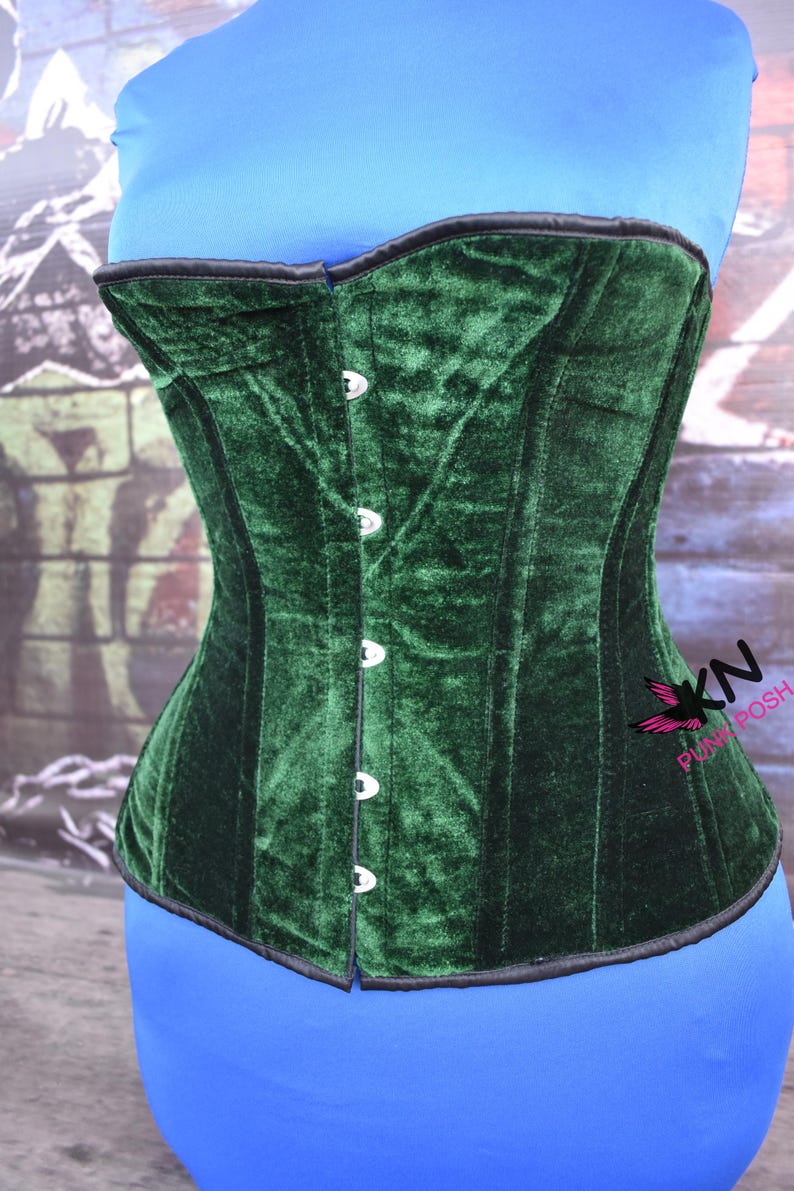 May include: A dark green velvet corset with a structured design. The corset features a row of silver-colored buttons down the front and black trim along the edges. The corset is displayed on a blue form.