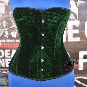 May include: A dark green velvet corset with a structured design. The corset has a black trim and silver-colored clasps down the front. The corset is displayed on a blue mannequin. The background features a collage of posters.