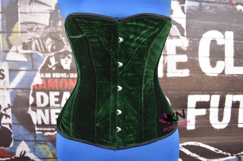 May include: A dark green velvet corset with a structured design. The corset has a black trim and silver-colored clasps down the front. The corset is displayed on a blue mannequin. The background features a collage of posters.
