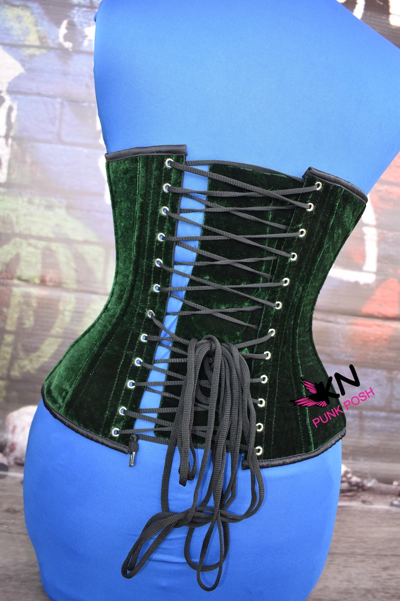 May include: A dark green velvet corset with black lacing. The corset is displayed on a blue form. The corset has a black trim and metal eyelets. The brand name "Punk Posh" is visible on the side.