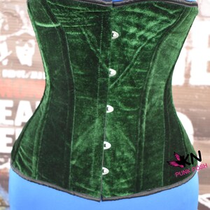 May include: A dark green velvet corset with a structured design. The corset features a row of silver-colored clasps down the front and a black trim along the top edge. The corset is displayed on a blue form.