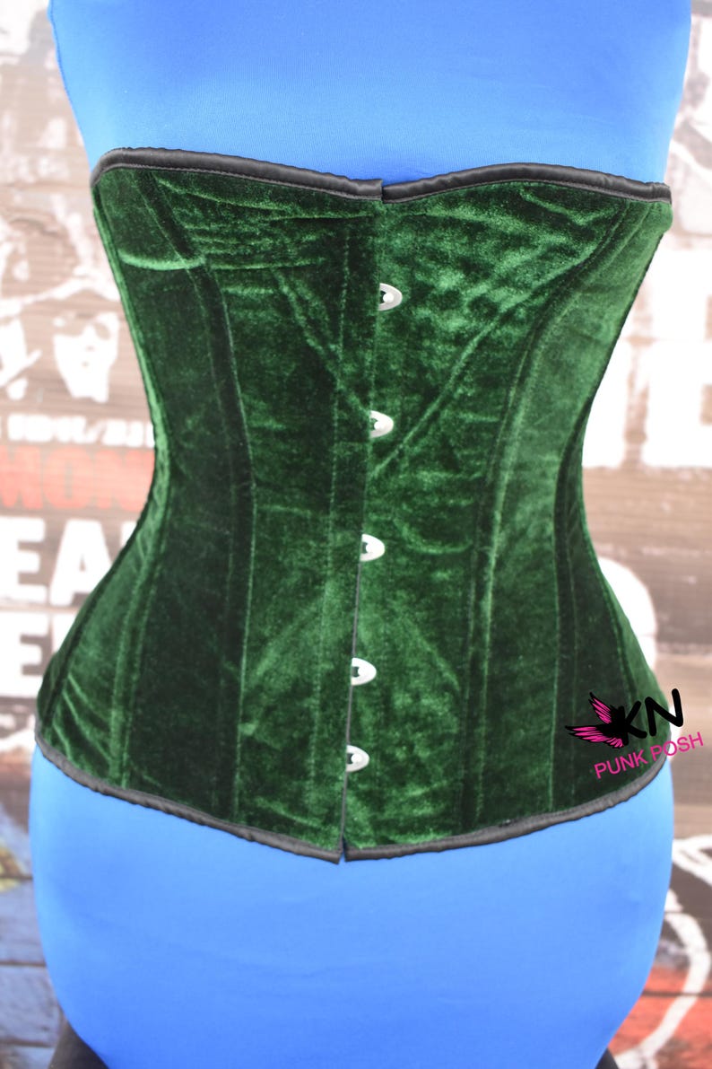 May include: A dark green velvet corset with a structured design. The corset features a row of silver-colored clasps down the front and a black trim along the top edge. The corset is displayed on a blue form.