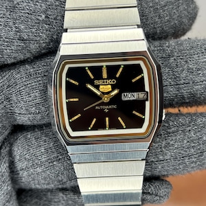 May include: A silver-toned Seiko automatic wristwatch with a black face. The watch features a rectangular face with gold accents, a day-date display, and a silver band. The watch face displays the day of the week and the date.