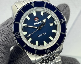 May include: A stainless steel Rado Captain Cook watch with a blue dial and bezel. The watch features white hour markers, a date window, and the text "Captain Cook". The watch is on a stainless steel bracelet.