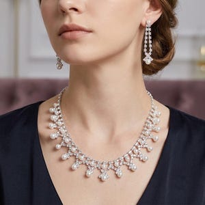 May include: A silver-toned necklace and matching drop earrings set featuring clear crystals and white faux pearls. The necklace has a scalloped design with dangling pearls, while the earrings consist of a vertical crystal chain ending in a pearl.