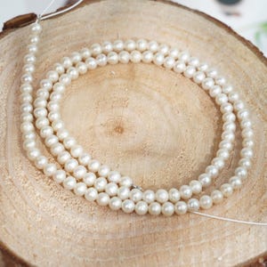 Peut inclure: Trois rangées de petites perles blanches et rondes sont disposées sur une surface en bois clair et texturé. Les perles sont enfilées sur un fin fil blanc, dont les extrémités sont visibles. Les perles ont un éclat subtil.