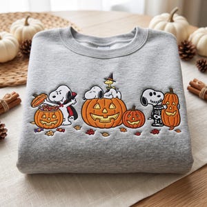 May include: A heather gray crewneck sweatshirt featuring an embroidered design of Snoopy and Woodstock celebrating Halloween. The embroidery depicts Snoopy dressed as a vampire, a skeleton, and resting on a large jack-o'-lantern with pumpkins.