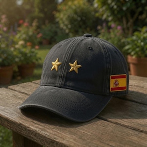 May include: A charcoal grey baseball cap featuring two embroidered gold stars on the front panels and a rectangular patch of the Spanish flag stitched onto the side. The cap has a curved brim and a structured crown with visible stitching details.