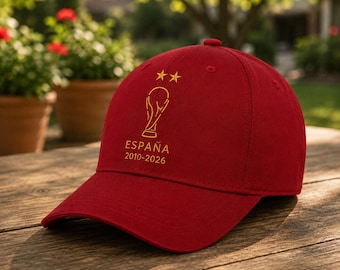 May include: A red baseball cap featuring an embroidered gold trophy icon, two gold stars above it, and the text ESPAÑA 2010-2026 printed in gold lettering on the front panel. The cap has a curved brim and a structured crown design.