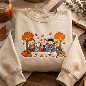 May include: A beige crewneck sweatshirt featuring an embroidered graphic of the Peanuts characters sitting on a wooden bench under autumn trees. The design includes Snoopy, Charlie Brown, and friends holding mugs, surrounded by orange pumpkins and leaves.