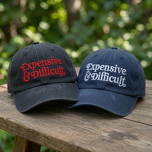 May include: Two baseball caps, one black and one navy blue, both featuring the embroidered text Expensive & Difficult. The black cap has red lettering, while the navy blue cap has white lettering. Each hat has a curved brim and a structured crown.