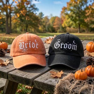 May include: An orange baseball cap with the word Bride embroidered in white, and a black baseball cap with the word Groom embroidered in white. Both caps feature small white embroidered stars, a crescent moon, and bat silhouettes.
