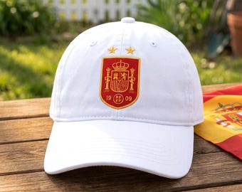 May include: A white baseball cap featuring an embroidered red and gold crest on the front panel. The crest displays a crown, pillars, and the text 19 RFEF 09, topped with two gold stars. The cap has a structured crown and a curved brim.