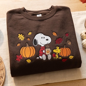 Könnte beinhalten: Ein dunkelbraunes Sweatshirt mit Rundhalsausschnitt und einer Stickerei von Snoopy, der einen roten Pullover trägt und eine Tasse hält, neben Woodstock sitzend. Das Motiv zeigt orangefarbene Kürbisse, bunte Herbstblätter und eine Eichel.