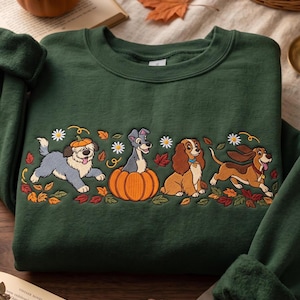 May include: A dark green crewneck sweatshirt featuring an embroidered design of four dogs surrounded by autumn leaves, white daisies, and pumpkins. The dogs include a fluffy grey dog with a pumpkin hat, a grey dog in a pumpkin, a brown spaniel, and a brown hound.