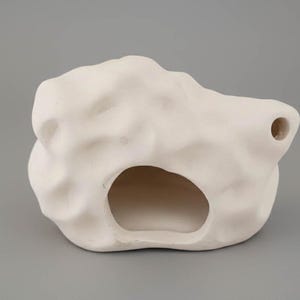 Puede incluir: Una escultura de cerámica abstracta de color crema, con una abertura en forma de cueva y un pequeño agujero. La superficie texturizada tiene un aspecto orgánico, similar a la roca. La escultura está sobre un fondo gris neutro.