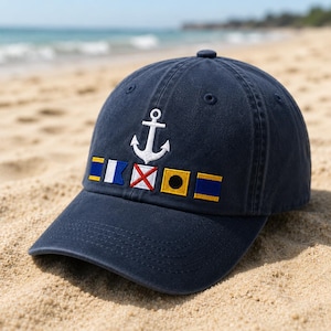 May include: A navy blue baseball cap featuring an embroidered white anchor centred above a horizontal row of five colourful nautical signal flags. The cap has a structured crown, a curved brim, and stitched eyelets on the top panels.