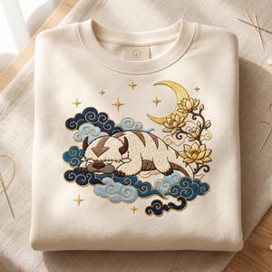 May include: A cream-colored crewneck sweatshirt featuring an embroidered design of a sleeping bison creature resting on blue and light blue stylized clouds. The scene includes a golden crescent moon, yellow lotus flowers, and small stars.