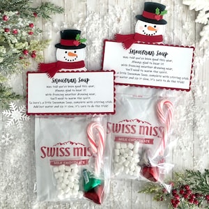 May include: A snowman-themed hot chocolate mix in a clear plastic bag with a red and white candy cane, white marshmallows, and chocolate candies. The bag has a red and white snowman design and the text "Snowman Soup" with instructions to add hot water and stir. The bag is labeled "Swiss Miss Milk Chocolate".