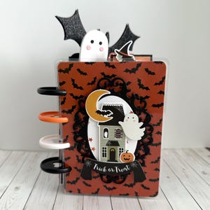 May include: A disc-bound notebook with an orange cover featuring a black bat pattern. The front displays a layered die-cut design with a haunted house, a crescent moon, a ghost, a pumpkin, and a banner with the text Trick or Treat.