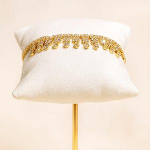 Puede incluir: Un collar dorado con un dise&ntilde;o brillante y tachonado de gemas. El collar se exhibe sobre un coj&iacute;n blanco, mostrando sus intrincados detalles y su elegante estilo. El collar presenta una serie de adornos colgantes en forma de l&aacute;grima.