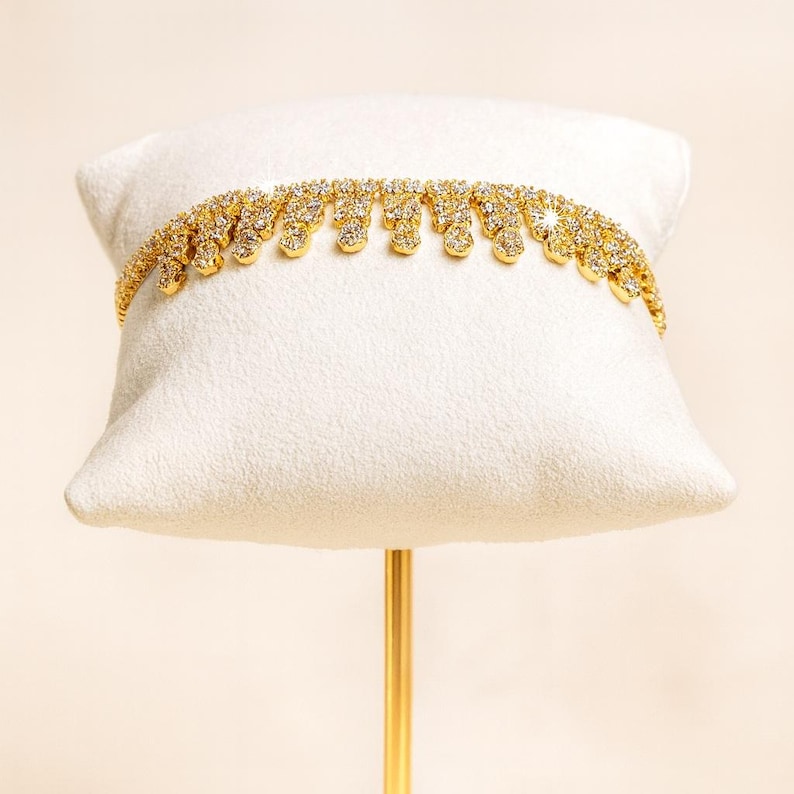 Puede incluir: Un collar dorado con un dise&ntilde;o brillante y tachonado de gemas. El collar se exhibe sobre un coj&iacute;n blanco, mostrando sus intrincados detalles y su elegante estilo. El collar presenta una serie de adornos colgantes en forma de l&aacute;grima.