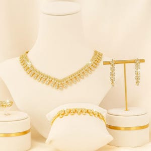 Puede incluir: Un conjunto de joyas doradas exhibido sobre soportes blancos. El conjunto incluye un collar, una pulsera, un anillo y pendientes colgantes. El collar y la pulsera presentan un patr&oacute;n repetido de peque&ntilde;as piedras brillantes.