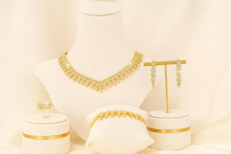 Puede incluir: Un conjunto de joyas doradas exhibido sobre soportes blancos. El conjunto incluye un collar, una pulsera, un anillo y pendientes colgantes. El collar y la pulsera presentan un patr&oacute;n repetido de peque&ntilde;as piedras brillantes.