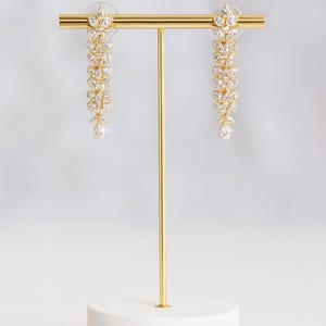 Puede incluir: Un par de pendientes colgantes dorados con dise&ntilde;o floral, exhibidos en un soporte dorado con base blanca. Cada pendiente presenta una cascada de cristales claros y brillantes, culminando en un cristal &uacute;nico y m&aacute;s grande.