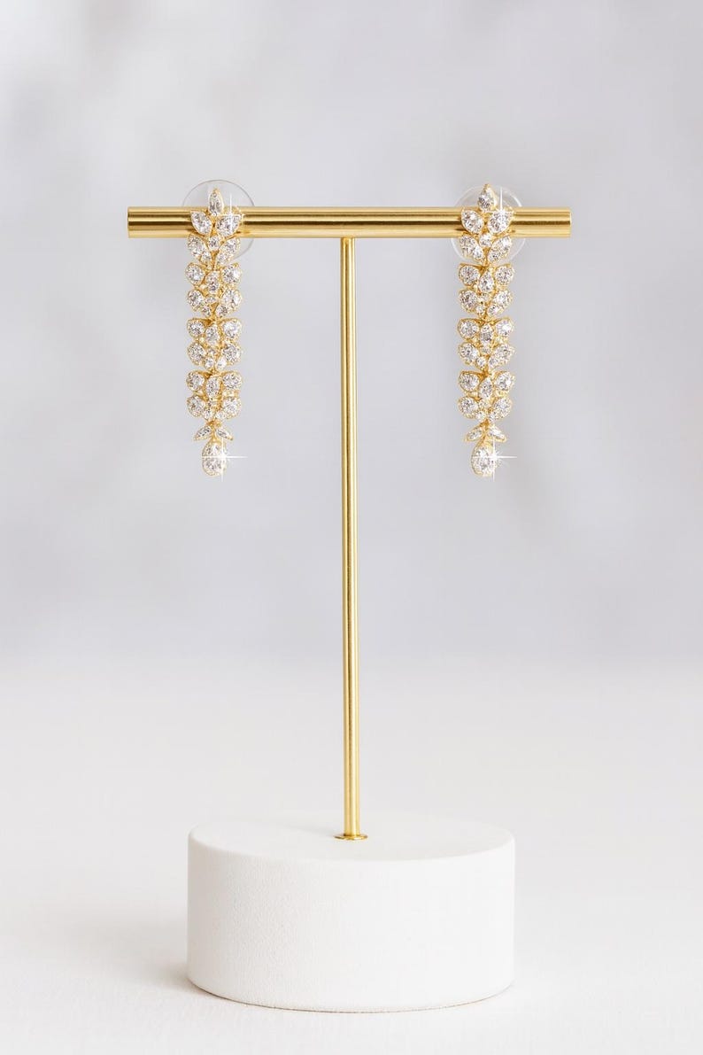 Puede incluir: Un par de pendientes colgantes dorados con dise&ntilde;o floral, exhibidos en un soporte dorado con base blanca. Cada pendiente presenta una cascada de cristales claros y brillantes, culminando en un cristal &uacute;nico y m&aacute;s grande.