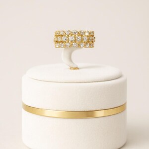 Puede incluir: Un anillo de oro con tres filas de piedras preciosas claras, exhibido en un soporte blanco. El anillo se encuentra sobre un fondo blanco, con una banda dorada alrededor de la base del soporte. Es una joya.