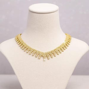 Puede incluir: Un collar dorado con un dise&ntilde;o delicado, exhibido en un busto de maniqu&iacute; blanco. El collar presenta una serie de peque&ntilde;os adornos brillantes y un colgante central. El dise&ntilde;o general es sim&eacute;trico y elegante.