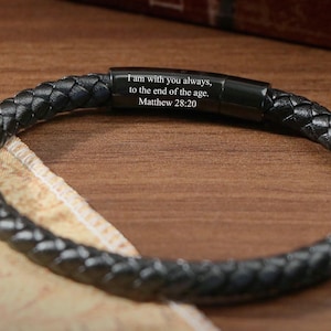 May include: Black braided leather bracelet with a magnetic clasp. The bracelet has the text "I am with you always, to the end of the age. Matthew 28:20" engraved on the clasp. The bracelet is displayed on a wooden surface.