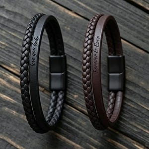 May include: Two braided leather bracelets with matte black metal clasps. The black bracelet is engraved with the text love you babe, and the dark brown bracelet is engraved with the text Forever & Ever. Both bracelets feature a dual-strand design.