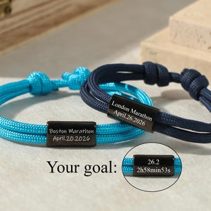 Könnte beinhalten: Zwei geflochtene Seilarmbänder, eines türkis und eines marineblau, mit schwarzen rechteckigen Anhängern. Die Anhänger sind mit "Boston Marathon April.20.2026" und "London Marathon April.26.2026" graviert. Ein dritter Anhänger lautet "26.2 2h58min53s".