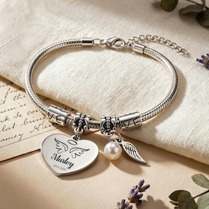 Könnte beinhalten: Ein silberfarbenes Schlangenkettenarmband mit einem herzförmigen Anhänger, der mit dem Namen Marley, den Daten 1952-2026 und Engelsflügeln graviert ist. Das Armband enthält zwei zylindrische Perlen mit dem Wort LOVE, eine weiße Perle und einen Flügelanhänger.