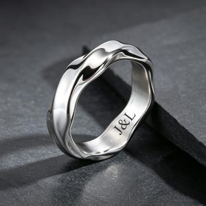 Op de afbeelding: Een gepolijste zilverkleurige ring met een golvend, onregelmatig bandontwerp. De binnenkant van de band is voorzien van de gegraveerde tekst J&L in een strak, donker lettertype. Het metalen oppervlak reflecteert het licht op de ring.