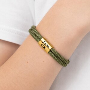May include: Olive green rope bracelet with a gold-colored clasp. The clasp features the name "Leon" and a fish symbol. The bracelet is worn on a wrist, and the background is a white shirt.
