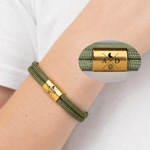 May include: A bracelet featuring two strands of olive green cord and a gold-toned metal cylindrical bead. The bead is engraved with a black design consisting of a crescent moon, a sun, and the letters A and D separated by crossed lines.