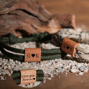 Puede incluir: Dos pulseras personalizadas con cuentas de madera. Una pulsera es verde oliva con las iniciales "G K" y un corazón. La otra es beige y bronceado con las iniciales "A L" y un corazón. La parte posterior de una pulsera dice "Love you forever".