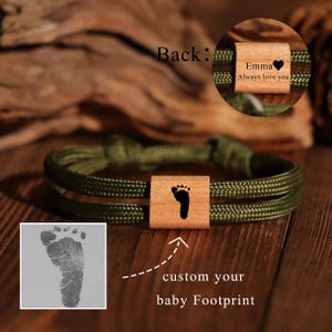 Könnte beinhalten: Olivgrünes Seilarmband mit einem Holzmittelstück mit einem Babyfußabdruck. Der Text "custom your baby Footprint" ist sichtbar. Eine Nahaufnahme zeigt den Text "Emma Always love you" auf der Rückseite.