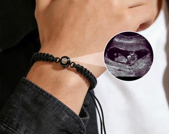 May include: Black braided bracelet with a circular charm and a small, round, dark-colored bead. An ultrasound image of a fetus is displayed next to the bracelet. The bracelet is worn on a wrist.