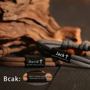 May include: Close-up of three braided rope bracelets. Two bracelets are brown and gray, with black rectangular clasps engraved with names and a cross. The third bracelet is brown with the text "FIRST COMMUNION September 06.2025".