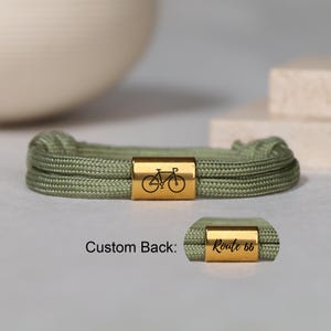May include: A sage green cord bracelet featuring a rectangular gold-toned metal bead engraved with a black bicycle icon. An inset shows the reverse side of the bead, which is engraved with the black cursive text Route 66 on a gold surface.