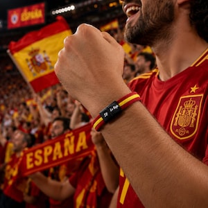 May include: A red, yellow, and black bracelet with the text "19 La Roja" on a black clasp. The bracelet is worn on a person's wrist. The person is wearing a red jersey with a yellow stripe and a Spanish coat of arms.