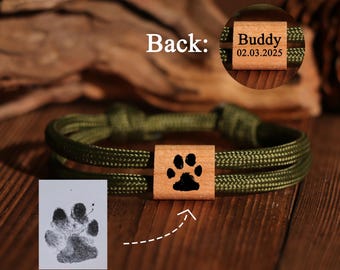 May include: Olive green rope bracelet with a wooden square featuring a black paw print. The back of the wooden square has the name "Buddy" and the date "02.03.2025" engraved. A paw print is shown on a white paper.
