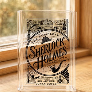 May include: A clear acrylic book-shaped display stand featuring black printed graphics. The design includes the text The Sherlock Holmes Museum, The Complete Sherlock Holmes Collection, and Sir Arthur Conan Doyle, with icons of a pipe, hat, and carriage.