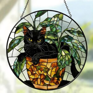 May include: Circular stained glass window art depicting a black cat in a plant pot. The pot is orange and yellow, and the leaves are green. The window is framed in black and suspended by a chain.