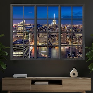 May include: A framed wall art piece depicting a nighttime cityscape viewed through a window. The image showcases a vibrant skyline with illuminated skyscrapers, a river, and a dark blue sky. The frame is black, and the artwork is displayed above a wooden cabinet.