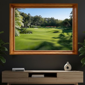 May include: A framed landscape painting of a golf course. The image features a lush green golf course with rolling hills, trees, and a pond. The frame is made of wood. The sky is blue with sunlight.